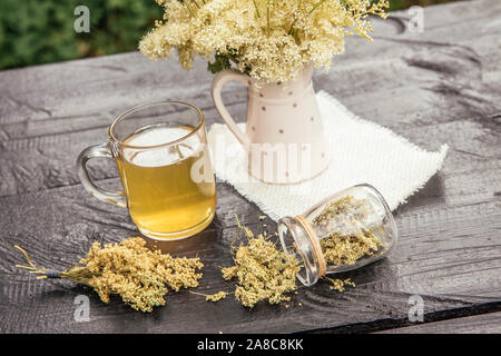 Fresh Meadowsweet, Filipendula ulmaria flowers in jug shape vase and it`s herbal tea in glass, tea powder in jar outdoors on garden table. Stock Photo